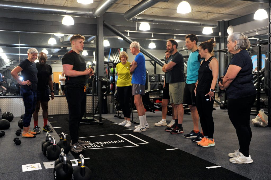 Nine gym members standing up listening to the gym instructor inside a gym, surrounded by dumbbells, kettle bells, medicine balls and a track ready for a circuit class.