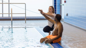 A swimming instructor crouching poolside next a senior man who is sitting with his legs in the water, holding his arm in the air, helping him stretch before he starts swimming.