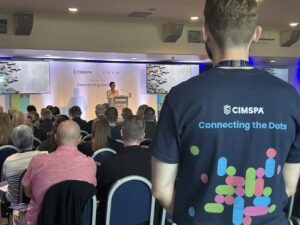 A man wearing a Connecting the Dots t-shirt faces away from the camera towards the stage where Tara Dillon is delivering a speech at CIMSPA conference 2025.